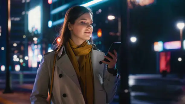 Photo of Portrait of a Beautiful Woman in Trench Coat Walking in a Modern City Street with Neon Lights at Night. Attractive Female Using Smartphone and Looking Around the Urban Cinematic Environment.