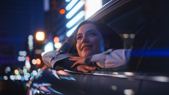 Una joven emocionada está sentada en el asiento trasero de un automóvil, viajando a casa por la noche. Mirando por la ventana con asombro de lo hermosa que es la calle de la ciudad con letreros de neón en funcionamiento. photo
