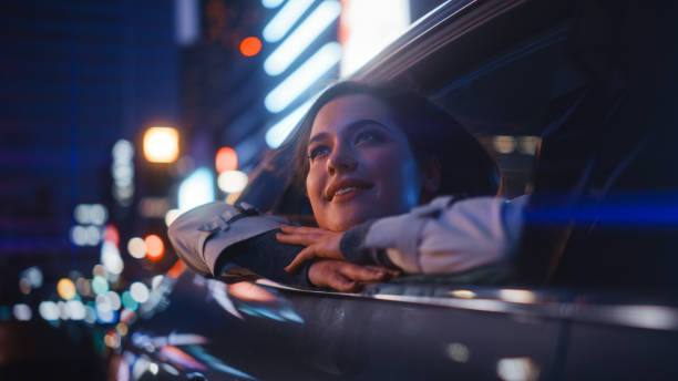 une jeune femme excitée est assise sur le siège arrière d’une voiture, rentrant chez elle la nuit. regarder par la fenêtre avec étonnement de la beauté de la rue de la ville avec des enseignes au néon qui fonctionnent. - night drive photos et images de collection