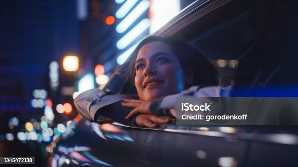 Excited Young Female Is Sitting On Backseat Of A Car Commuting Home At Night Looking Out Of The Window With Amazement Of How Beautiful Is The City Street With Working Neon Signs Stock Photo - Download Image Now