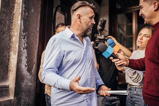 Group of people, male politician confronted by journalists with microphones.