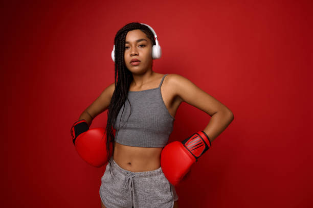 entre em contato com o conceito de arte marcial. jovem mulher esportiva boxeadora em fones de ouvido sem fio e roupas esportivas, coloca as mãos com luvas de boxe na cintura e olha para a câmera posando sobre fundo vermelho com espaço de cópia. - muay thai audio - fotografias e filmes do acervo