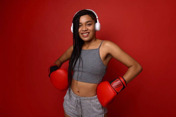 atleta mulher africana boxeadora em fones de ouvido sem fio e mãos em luvas de boxe na cintura, sorri sorriso dentuço olhando para a câmera, posando sobre fundo vermelho com espaço de cópia. entre em contato com o conceito de arte marcial. - muay thai audio - fotografias e filmes do acervo