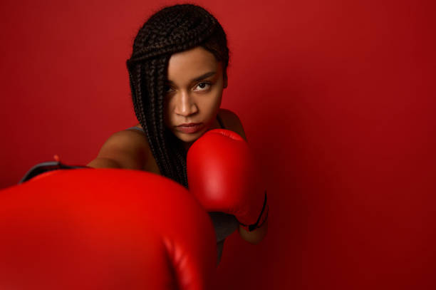 retrato de perto de jovem africana concentrada boxeadora usando luvas vermelhas de boxe, fazendo batidas diretas, isolada sobre fundo vermelho com espaço de cópia. mulher negra boxeador socando em direção à câmera - muay thai audio - fotografias e filmes do acervo