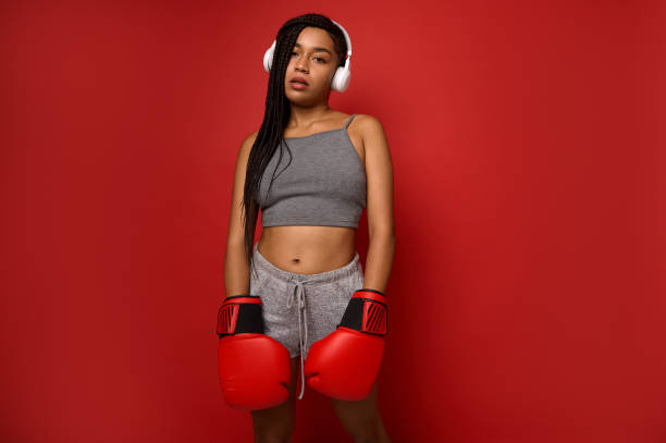 jovem atleta exausta boxeadora, esportista usando fones de ouvido sem fio, olha para a câmera posando com luvas vermelhas de boxe contra um fundo vermelho com espaço de cópia para anúncio. contato conceito de arte marcial - muay thai audio - fotografias e filmes do acervo