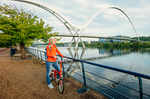 Mature woman on the promenade early morning with her bicycle pausing to view the Infinity Bridge at Stockton on Tees, England.