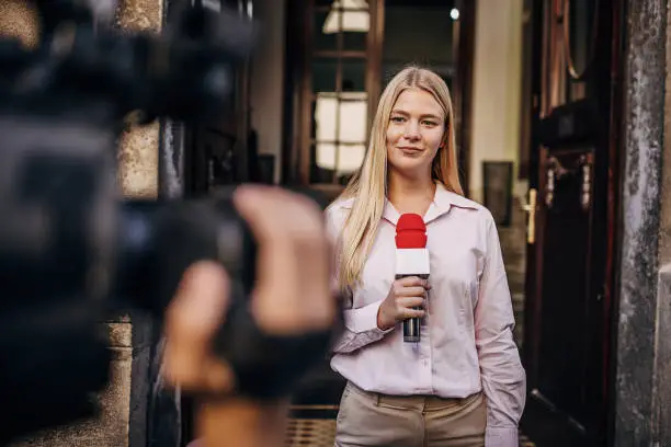 Two people, female television reporter holding a microphone, she is standing outdoors in front of a building. Male camera operator is filming her,
