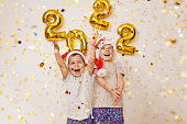 Happy smiling Kids in red Santa hat are enjoying confetti near white wall with gold balloons. Celebrating New Year 2022 at home. Christmas concept