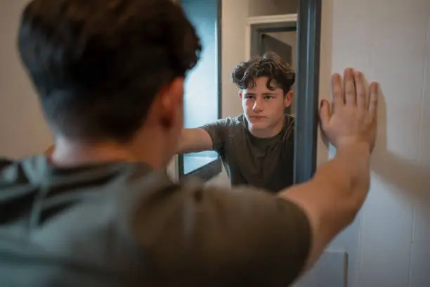 An over-the-shoulder shot of a young teenage male athlete in the gym changing rooms, he is taking a break from his gym session to catch his breath, he has his hands on the wall and is looking at his reflection in the mirror.