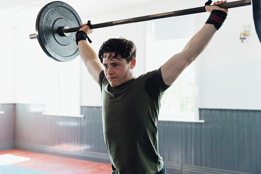 A close-up waist-up shot of a young teenage male athlete lifting his weights, he is performing an overhead press with a barbell.