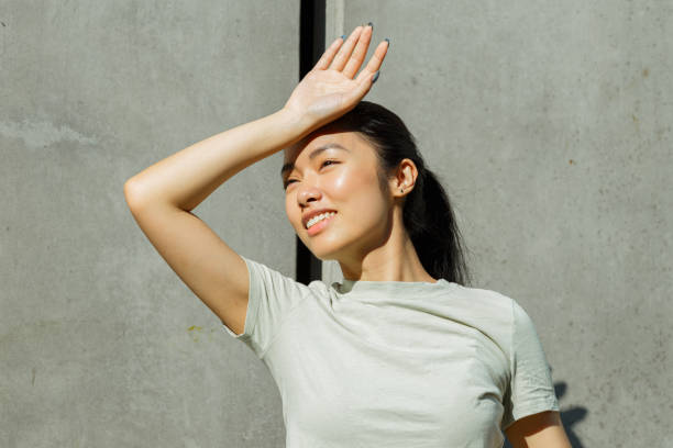 Asian sportswoman covers eyes from sun on sunny city street Happy young Asian sportswoman covers eyes from sun with palm standing near wall with large concrete plates on sunny city street hot vietnamese women pictures stock pictures, royalty-free photos & images