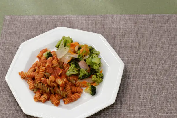 Prepared meal of vegetable chickpea pasta with meat tomato sauce, broccoli, onions, carrots and black pepper on a plate. Meal of rotini pasta with meat sauce and vegetables on a white plate close-up.