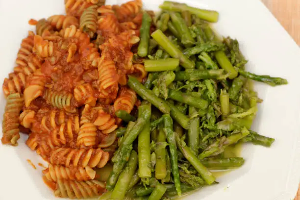 Close-up of prepared food meal of vegetable rotini pasta with tomato meat sauce with green asparagus on the side. Cooked asparagus with butter alongside chickpea vegetable rotini pasta and meat sauce.