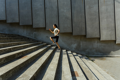 Long haired Asian woman in tracksuit runs up stone stairs past wall with concrete panels training on city street side view
