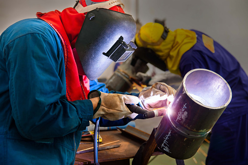 The welder is welding the steel pipe with Tungsten Inert Gas Welding process (TIG). The welder wears protective equipment with a mask and heat resistant gloves.