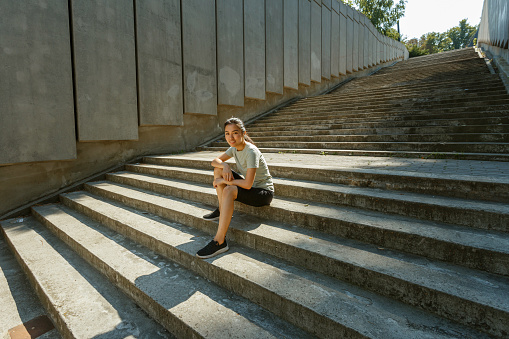 Attractive Asian woman in tracksuit and sneakers sits resting on empty grey steps casting shadow after outdoor training in sunny city