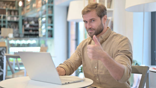 istock Thumbs Up by Young Man while Working on Laptop in Cafe 1345510499