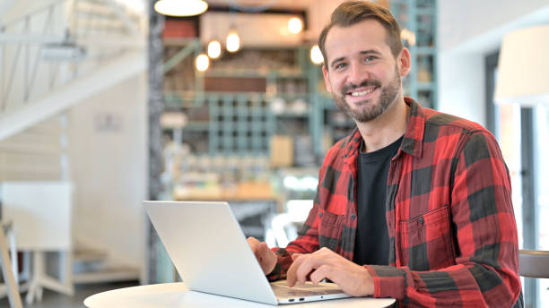 istock Beard Young Man with Laptop Smiling at Camera in Cafe 1345510921