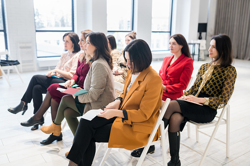 Group of females sitting in the audience for business training. Women outlining coach speech in notebook. People attending personal development seminar or conference. Lifelong learning concept