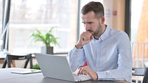 istock Sick Young Man with Laptop Coughing in Office 1345515400