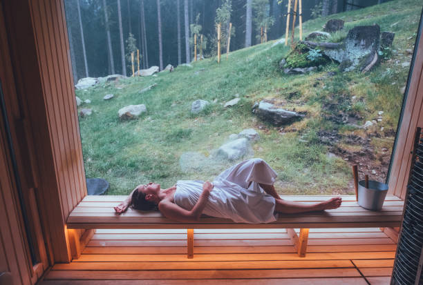 Relaxing Woman wrapped white towel lying on the wooden bench in Hot Finnish sauna with a huge wide window with green forest view and enjoying pleasant healthy body care temperature treatment. stock photo