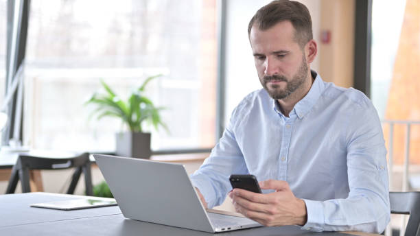 istock Young Man using Laptop and Smartphone in Office 1345515703