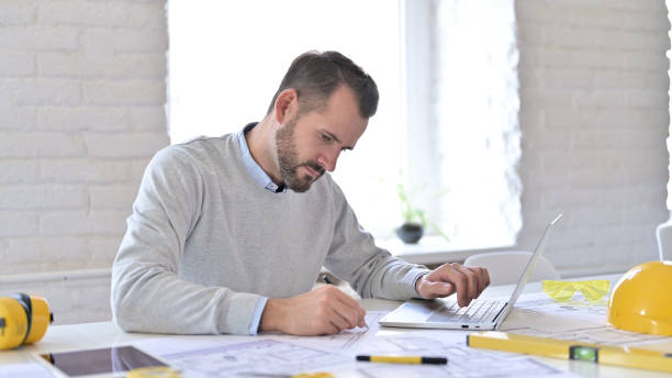 istock Young Architect with Laptop doing Paperwork in Office 1345518118