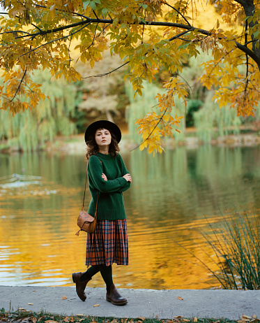 Young Caucasian woman in green sweater walking near the lake in park in autumn