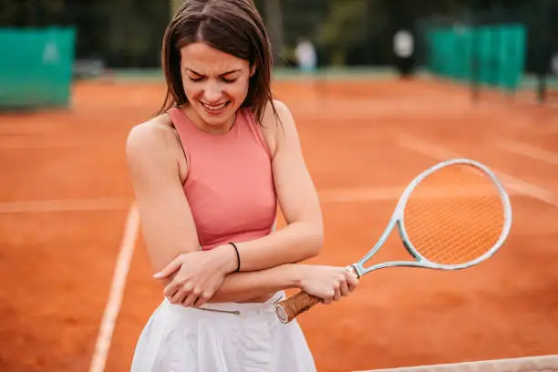 Sporty young woman holding her elbow in pain while playing tennis on a tennis court.