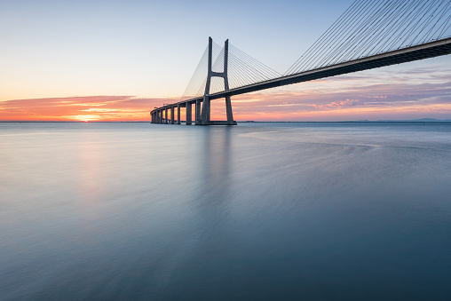 The Zeelandbrug during sunrise. The sun is already a bit higher in the sky and it’s reflected in the water of the North sea. The bridge is also reflected in the water. Cars drive at a high speed over it.