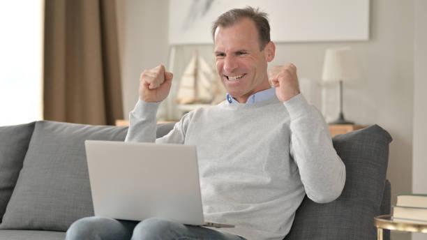 istock Surprised Middle Aged Businessman Celebrating Success on Laptop on Sofa 1345525901