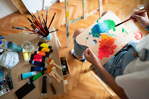 Unrecognizable female artist, sitting in front of an easel, mixing acrylic paint on wooden artist's palette