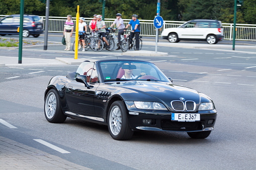 Adult man is driving BMW Z 3 roadster over intersection of B224 in Essen Werden. In background a a group of people and cyclists is waiting at stoplight. Some traffic is behind.