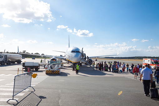 Queue of people entering Ryanair airplane at airport Weeze in summertime. People walk to airplane from terminal. Airport staff is standing in front of airplane. At right side ambulance is standing. At left side is tanker truck