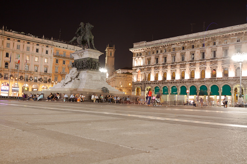 Square Piazza del Duomo with monument of Vittorio Emanuele II in Milan at night. Long exposure. People are sitting around monument. At right side is Galleria Vittorio Emanuele II