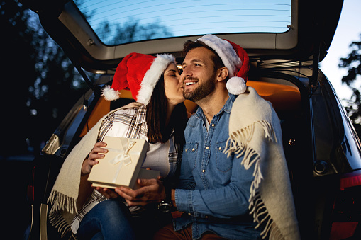Young couple in love sitting in car open trunk on Christmas exchanging gifts