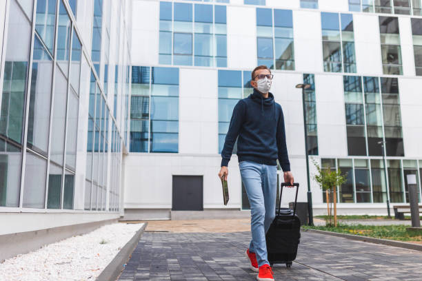 istock Young man with glasses walking outside an airport while wearing a protective mask and dragging his suitcase 1345555830