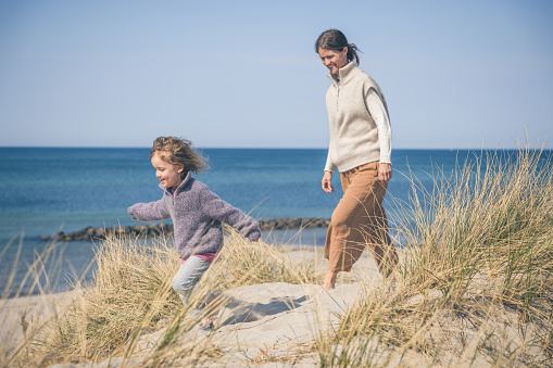 Mother and cute little danish girl with blond hair wearing beautiful homemade knitwear while having fun outdoors in nature while playing on sand dunes on beach