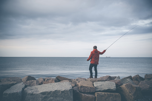 Saltwater Fly-fishing from a rocky pier in the capital city Copenhagen, Denmark during autumn, winter or fall.  Male angler wearing fashionable red  jacket and knit hat.
