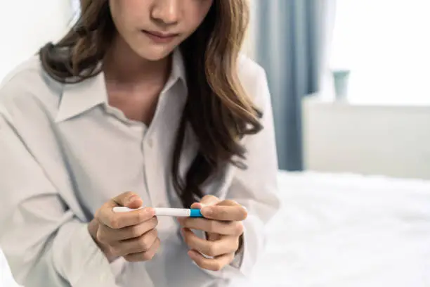 Photo of Close up of woman feeling shock after look at positive Pregnancy test. Disappointed attractive beautiful unplanned pregnancy girl sit on bed with upset and sad face for problem don't want to pregnant.
