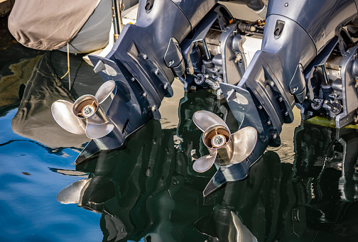 Close up view of the stern of a boat with two outboard motors that are up and out of the water, their props are just above the water.  The water is calm and the props are being reflected in the water.