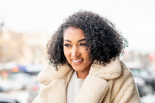 A beautiful woman of African decent, with flawless skin and sparkling white teeth, stops to pose for a portrait in the street.  She is dressed warmly in a cream fleece coat and has  warm smile on her face.