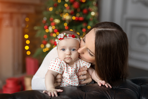 A loving mother kisses her child on Christmas Day in a room with Christmas decorations, a Christmas tree, lights and gifts. Love and care of parents