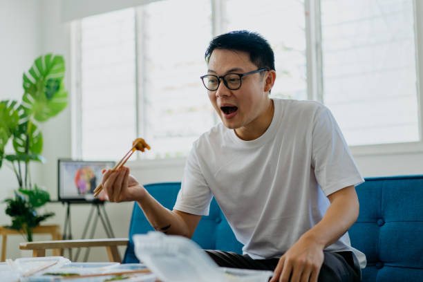 Asian man eating home delivery meal in living room Image of an Asian Chinese man eating home delivery meal in living room Man EATING FOOD stock pictures, royalty-free photos & images