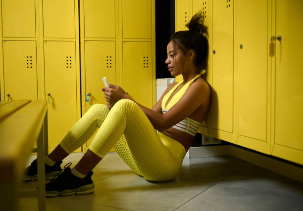 Young beautiful African female athlete, sports woman dressed in a yellow tight-fitting tracksuit, sits on the floor in the locker room and swiping in mobile phone. Sporty woman relaxing after workout Young beautiful African female athlete, sports woman dressed in a yellow tight-fitting tracksuit, rests after hard workout, sits on the floor in the gym locker room and swiping in mobile phone. women boxing sport exercising stock pictures, royalty-free photos & images