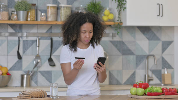 キッチンでスマートフォンでオンラインショッピングをしているアフリカの女性 - 24120 ストックフォトと画像