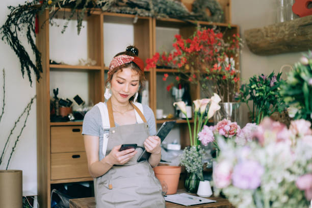 junge asiatische floristin, inhaberin eines blumenladens für kleine unternehmen, mit digitalem tablet und smartphone am arbeitsplatz. lagerbestände prüfen, kundenbestellungen entgegennehmen, produkte online verkaufen. tägliche routine, ein kleines unte - order buying purchase order bill stock-fotos und bilder