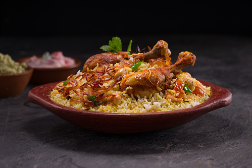 Chicken dhum biriyani using jeera rice and spices arranged in  earthen ware with raitha and lemon pickle  on grey background.