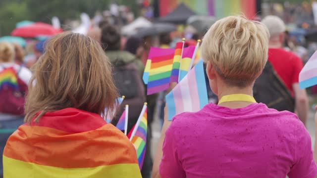 Two women watch a pride parade, one wearing a rainbow flag around her shoulders the blonde is a bright pink top and waving small diverse flags denoting gays, bisexuals and transgenders, supporting human rights