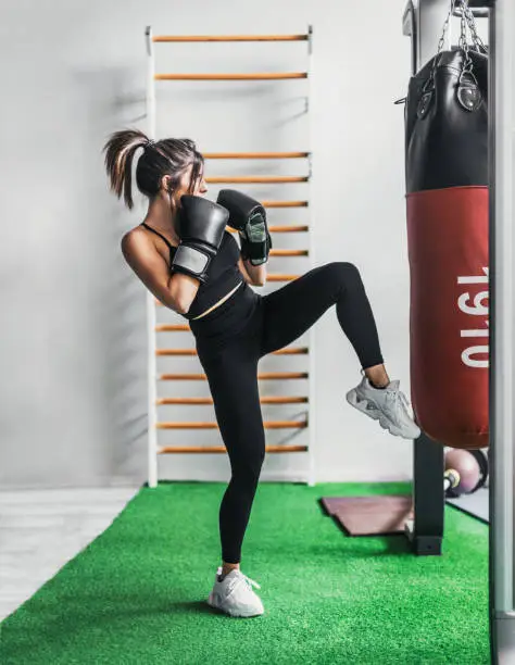 a full-bodied girl wearing black boxing gloves kicks a training bag.